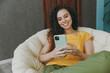 © ViDi Studio - Young smiling happy woman of African American ethnicity wear casual clothes use mobile cell phone sits in armchair stay at home flat rest relax spend free spare time in living room indoors grey wall.