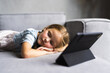 © F8  \ Suport Ukraine - Smiling little girl lying on comfortable couch, enjoying playing online game on digital tablet computer.