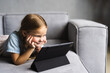 © F8  \ Suport Ukraine - My favorite pastime. Smiling small child girl sit on comfy sofa in modern studio apartment hold tablet computer read book online use handy web app. Little kid rest on cozy couch play videogames on pad