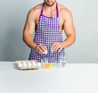 © oleg_ermak - Chef bodybuilder in protective apron and cook eggs on whie kitchen table. Muscular man cooking. Healthy food and dieting. Food additives for sportsman, daily calorie diet