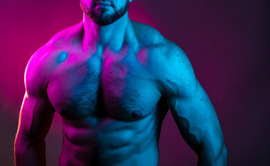 Sportive man bodybuilder is posing in the colorful neon light with naked muscular torso showing chest, abdominal muscles in neon studio light.