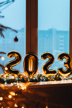 2023 Golden Foil Balloons On Blue Window Sill. Celebrating Holidays At Home, Festive Decor Concept. Happy New Year 2023. Close-up Numbers Of Year 2023 On Dark Background. Bokeh Warm Garland Light.