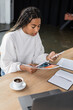 © LIGHTFIELD STUDIOS - African american businesswoman using smartphone and holding paper with charts near coffee in office.