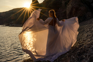 A mysterious female silhouette with long braids stands on the sea beach with mountain views, Sunset rays shine on a woman. Throws up a long white dress, a divine sunset.