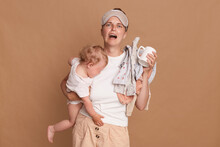 Horizontal Shot Of Desperate Sad Frustrated Mother With Baby Daughter And Laundry In Hands, Being Tired, Needs Rest, Crying, Expressing Sorrow, Posing Isolated Over Brown Background.