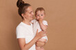 © sementsova321 - Portrait of happy delighted dark haired woman with bung hairstyle standing with baby daughter in hands, mother looking at her child with love and smile isolated over brown background.