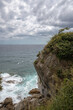 Vertical photo shows a rocky with vegetation. Wavy sea and sky with clouds at he background.