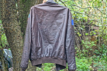 one old military aviation brown leather pilot jacket hangs on a wire among the green vegetation on the street