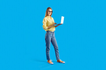 Happy young woman using modern laptop computer. Full body studio shot of beautiful student girl in shirt, jeans and eyeglasses standing against solid blue background, holding notebook PC and smiling