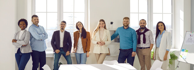 Portrait of friendly and joyful startup colleagues working together in bright office in modern business center. Smart multiracial men and women stand together in row and smile while looking at camera.