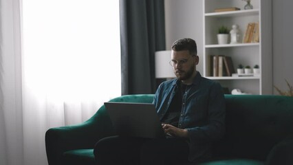 Bearded young man in denim shirt working on laptop, handsome coder writing code sitting on the couch