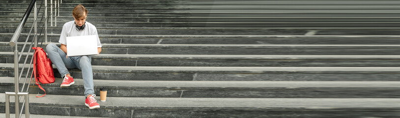 Guy student with laptop sits on the steps outdoors. Selective focus, banner