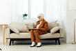 © Tatiana - a full-length photo of a nice sweet old lady sitting on a beige sofa in a brown suit working on a laptop