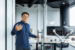 © Liubomir - A successful engineer programmer boss invites to his office, an Asian man in a shirt and glasses looks at the camera and smiles and waves to the interlocutor.