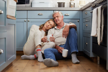 Senior couple, love and support while sitting together feeling safe, relax and calm drinking coffee and bonding comfortable on a kitchen floor. Happy old man and woman enjoying retirement together