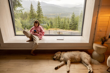 woman sits with mobile phone on window with great view on mountains behind while resting with her do