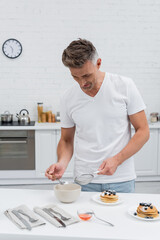 Canvas Print - Man taking powdered sugar from bowl and holding sieve near pancakes in kitchen.