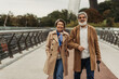 © LIGHTFIELD STUDIOS - cheerful senior woman walking with happy husband holding umbrella on bridge.