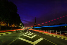 A View Along A City Road At Night, Tall Buildings And Light Trails, The Eiffel Tower In The Distance.