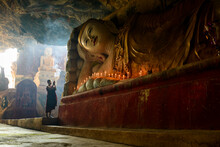 A Cave Temple, A Large Reclining Buddha Statue, Lit Candles And A Monk Praying.