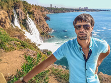 Young Adult Male Tourist Posing In Front Of Lower Duden Waterfall In Antalya, Turkey. A Man In Sunglasses And A Blue Shirt Stands Against A Blurred Seascape