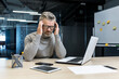 © Liubomir - Tension headache at work. A young handsome gray-haired male businessman in glasses sits in the office at a desk with a laptop and a phone, feels a headache, holds his head with his hands.