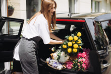 A Woman Florist Puts Flowers In The Trunk Of A Car. Flower Delivery Concept.