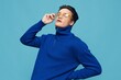 © Tatiana - a cute man stands on a light blue background in a blue zip-up sweater and holds yellow sunglasses with his hand, closing his eyes with pleasure