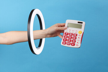 Wall Mural - A woman's hand holds a calculator through a led ring lamp on a blue background. Creative idea. Business, economy concept