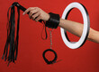 Woman's hand in sex shop leather handcuffs holding whip through led ring lamp on red background. Sex games. Creative idea.