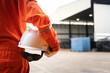 An operation worker in orange coverall is holding white safety helmet or hardhat with factory place as background. Safe working practice in the industrial scene photo, close-up and selective focus.