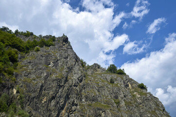 View from below of high rocky mountain with abundant greenery against blue sky with clouds. Selective focus.