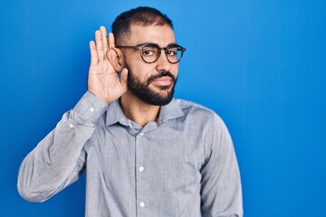 Canvas Print - Middle east man with beard standing over blue background smiling with hand over ear listening an hearing to rumor or gossip. deafness concept.