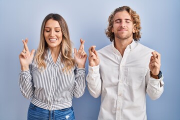 Poster - Young couple standing over blue background gesturing finger crossed smiling with hope and eyes closed. luck and superstitious concept.