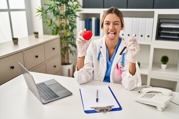 Poster - Young blonde woman wearing doctor uniform holding blood and heart sticking tongue out happy with funny expression.