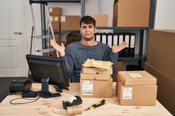 Poster - Young hispanic man working at small business ecommerce using smartphone clueless and confused expression with arms and hands raised. doubt concept.