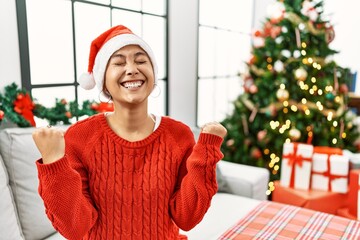 Wall Mural - Young hispanic woman with short hair wearing christmas hat sitting on the sofa celebrating surprised and amazed for success with arms raised and eyes closed. winner concept.