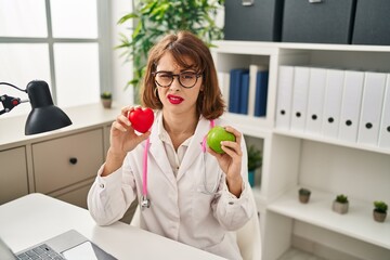 Poster - Young doctor woman holding heart and green apple clueless and confused expression. doubt concept.