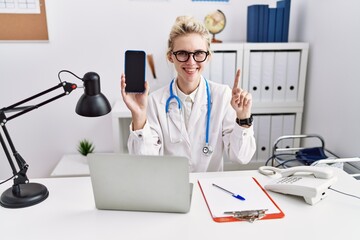 Sticker - Young doctor woman holding smartphone showing blank screen smiling with an idea or question pointing finger with happy face, number one