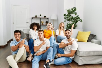 Canvas Print - Group of people sitting on the sofa and floor at home smiling and looking at the camera pointing with two hands and fingers to the side.