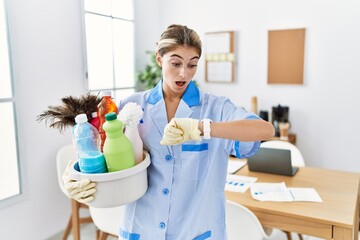 Poster - Young blonde woman wearing cleaner uniform holding cleaning products looking at the watch time worried, afraid of getting late