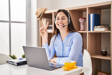 Poster - Young brunette woman wearing call center agent headset smiling with happy face winking at the camera doing victory sign. number two.