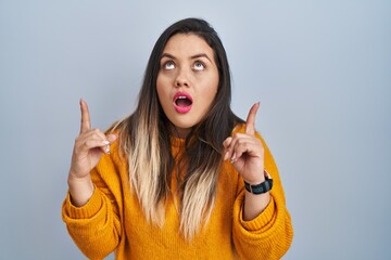 Wall Mural - Young hispanic woman standing over isolated background amazed and surprised looking up and pointing with fingers and raised arms.