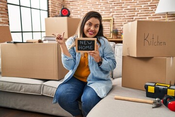 Wall Mural - Young hispanic woman holding blackboard with new home text smiling happy pointing with hand and finger to the side