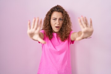 Wall Mural - Young caucasian woman standing over pink background doing stop gesture with hands palms, angry and frustration expression