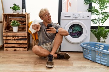 Wall Mural - Young blond man doing laundry using smartphone pointing to you and the camera with fingers, smiling positive and cheerful