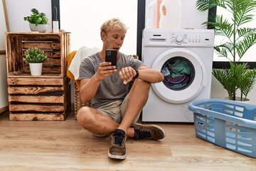 Poster - Young blond man doing laundry using smartphone looking at the watch time worried, afraid of getting late