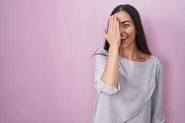 Poster - Young brunette woman standing over pink background covering one eye with hand, confident smile on face and surprise emotion.