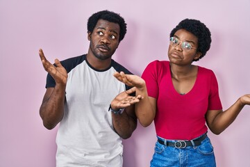 Poster - Young african american couple standing over pink background clueless and confused expression with arms and hands raised. doubt concept.