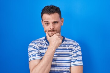 Poster - Young hispanic man standing over blue background looking confident at the camera smiling with crossed arms and hand raised on chin. thinking positive.